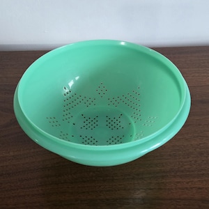 May include: A light green plastic colander with a wide rim and numerous small holes for draining. The colander is round and sits on a slightly raised base. The interior has a series of decorative hole patterns.