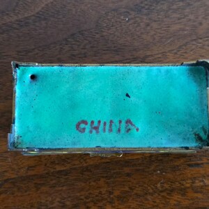 Puede incluir: Caja rectangular para baratijas con interior de esmalte turquesa y marco de metal dorado. La palabra "CHINA" est&aacute; impresa en rojo en el interior. La caja se muestra sobre una superficie de madera.