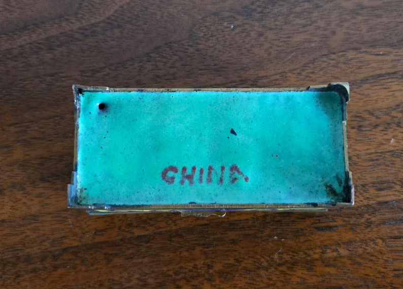 Puede incluir: Caja rectangular para baratijas con interior de esmalte turquesa y marco de metal dorado. La palabra "CHINA" est&aacute; impresa en rojo en el interior. La caja se muestra sobre una superficie de madera.