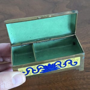 Puede incluir: Peque&ntilde;a caja de lat&oacute;n abierta con interior verde y un dise&ntilde;o floral decorativo amarillo y azul en la parte delantera. La caja es rectangular con tapa con bisagras y un peque&ntilde;o compartimento en el interior.