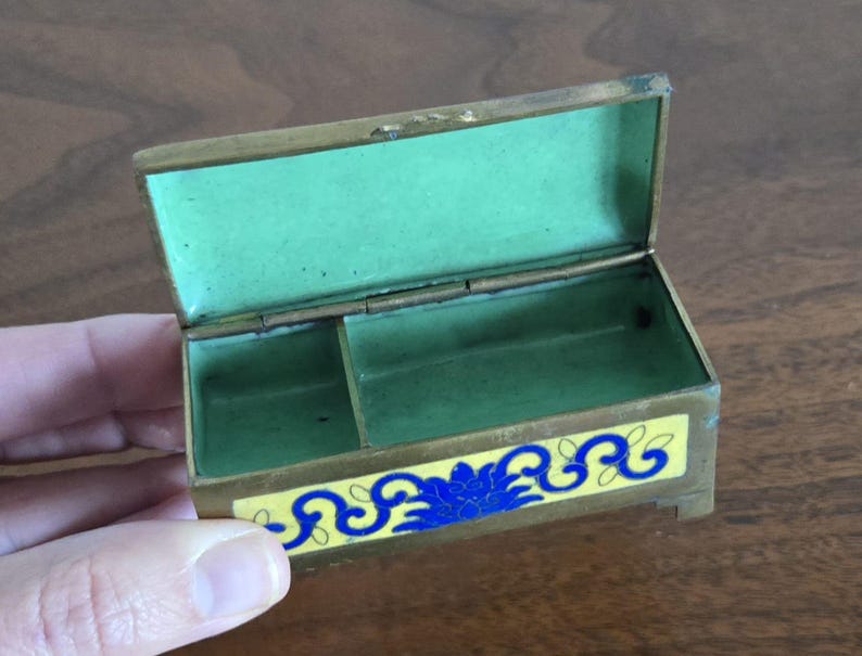 Puede incluir: Peque&ntilde;a caja de lat&oacute;n abierta con interior verde y un dise&ntilde;o floral decorativo amarillo y azul en la parte delantera. La caja es rectangular con tapa con bisagras y un peque&ntilde;o compartimento en el interior.