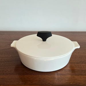 May include: A round, white ceramic casserole dish with a matching lid. The lid features a distinct, black, T-shaped handle centered on top. The dish has two small, integrated side handles for lifting, and the entire piece has a smooth, glossy finish.