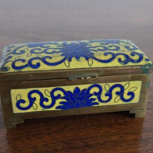 Puede incluir: Una caja de joyas rectangular antigua con marco y patas de lat&oacute;n. La caja est&aacute; decorada con esmalte amarillo y dise&ntilde;os florales azules. La tapa y los lados presentan dise&ntilde;os intrincados, creando una pieza de almacenamiento decorativa.