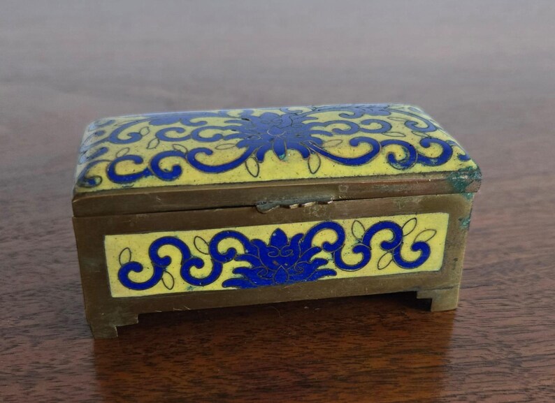 Puede incluir: Una caja de joyas rectangular antigua con marco y patas de lat&oacute;n. La caja est&aacute; decorada con esmalte amarillo y dise&ntilde;os florales azules. La tapa y los lados presentan dise&ntilde;os intrincados, creando una pieza de almacenamiento decorativa.
