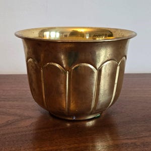 May include: A vintage brass planter with a scalloped design. The planter has a wide, rounded rim and a series of vertical, curved indentations around the body. The brass has a warm, golden hue and shows signs of age.