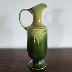 May include: A tall ceramic pitcher featuring a glossy finish with a gradient of deep forest green at the base transitioning into a lighter olive and yellow glaze near the top. The vessel has a curved handle, a flared rim, and a pedestal base.
