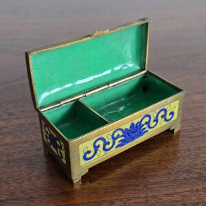 Puede incluir: Una peque&ntilde;a caja rectangular de lat&oacute;n con tapa con bisagras, abierta para revelar un interior verde brillante. El exterior presenta una base de esmalte amarillo con dise&ntilde;os florales azules. La caja est&aacute; elevada sobre peque&ntilde;os pies y tiene un borde dorado.