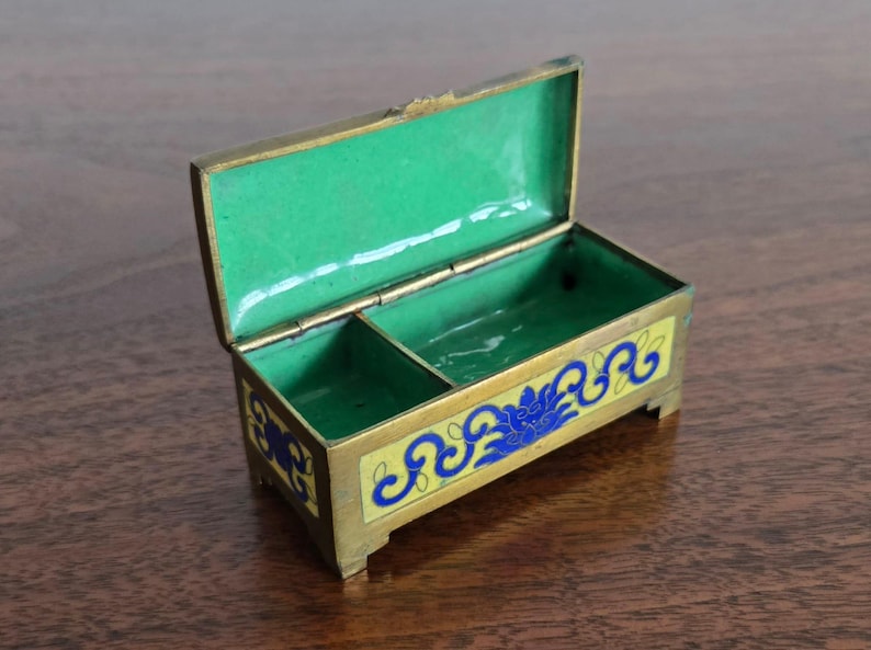 Puede incluir: Una peque&ntilde;a caja rectangular de lat&oacute;n con tapa con bisagras, abierta para revelar un interior verde brillante. El exterior presenta una base de esmalte amarillo con dise&ntilde;os florales azules. La caja est&aacute; elevada sobre peque&ntilde;os pies y tiene un borde dorado.