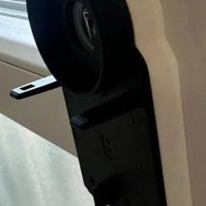 Könnte beinhalten: Ein schwarzer Fensterrollladenmechanismus aus Kunststoff mit einer weißen Kunststoffhalterung. Der Mechanismus hat eine runde, schwarze Rolle und einen weißen Kunststoffhebel.