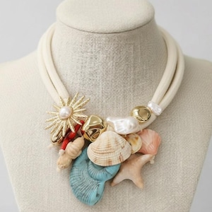 Op de afbeelding: Een crèmekleurige touwketting versierd met schelpen, parels en gouden accenten. De ketting heeft een gouden zonnebloemhanger met een parel in het midden en diverse schelpen in blauwe, roze en beige tinten.