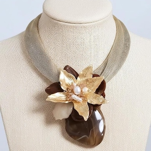 May include: A statement necklace with a gold-toned flower pendant, pearl accents, and a large brown stone. The pendant is attached to a mesh-like, gold-coloured collar. The necklace is displayed on a mannequin.