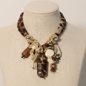 Puede incluir: Un collar llamativo con un cuello de tela con estampado de leopardo. El collar presenta detalles metálicos dorados, varias cuentas marrones y crema, y un colgante rectangular grande con un diseño de estampado de leopardo. También se incluye una perla.