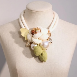 May include: Cream statement necklace with a large floral pendant. The pendant features cream fabric petals, pearls, and green leaf accents. The necklace is displayed on a mannequin. The necklace is approximately 40 cm in length.