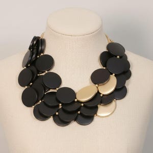 May include: A statement necklace featuring layered black and gold-tone circular pendants. The necklace is displayed on a mannequin, showcasing its design and structure. The necklace has a gold-tone chain.