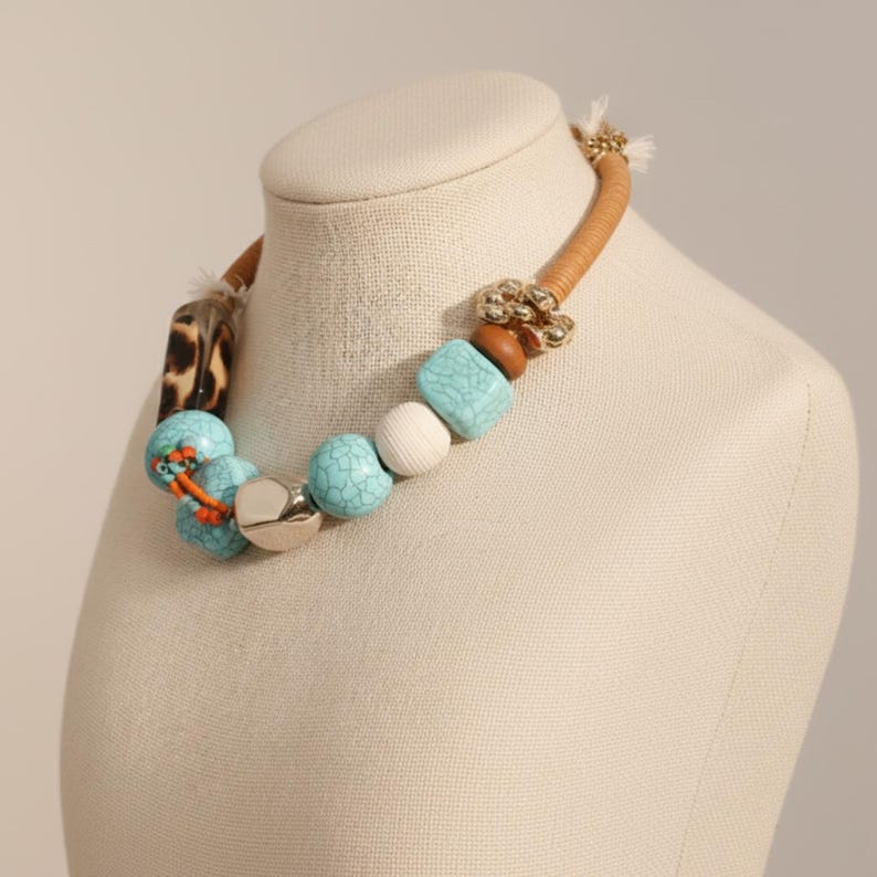May include: A statement necklace featuring a tan leather cord with a variety of beads. The beads include turquoise, white, and brown elements, along with a silver-toned accent and a leopard print section. The necklace is displayed on a mannequin.