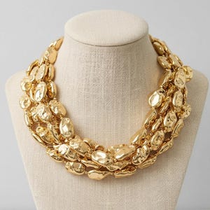 May include: A gold statement necklace with multiple strands. The necklace features irregularly shaped, textured metal pieces. The jewelry is displayed on a beige mannequin bust, showcasing its design and form.