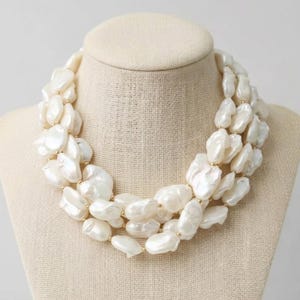 Puede incluir: Collar de perlas de tres hilos exhibido en un maniquí. El collar presenta grandes perlas blancas irregulares con detalles dorados. Las perlas están ensartadas para crear un efecto en capas, mostrando un diseño de joyería clásico.