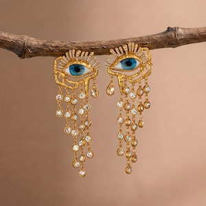 Può includere: Un paio di orecchini color oro con un motivo a occhio caratterizzato da iride e pupilla blu. La parte superiore è decorata con piccoli cristalli trasparenti, mentre diverse catenelle dorate con cristalli pendenti scendono verso il basso.