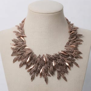 May include: A rose gold tone statement necklace featuring a dense arrangement of overlapping leaf-shaped metal components. Each leaf is textured with a dark, granular finish and interspersed with smooth, polished rose gold accents throughout.
