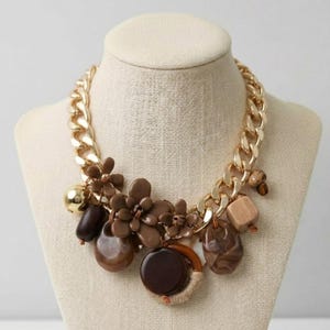 Puede incluir: Un collar de cadena dorada con varios abalorios marrones y beige. Los abalorios incluyen formas de flores, cuentas y formas geométricas. El collar se exhibe en un maniquí.
