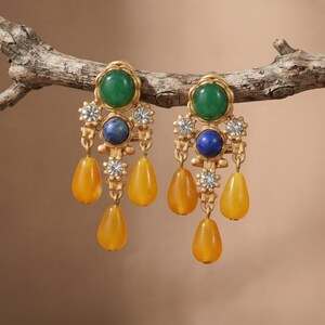 May include: A pair of gold-tone chandelier earrings featuring a round green cabochon at the top, a smaller round blue cabochon below, and three dangling yellow teardrop-shaped beads. The metal frame is accented with small, clear, round rhinestones.