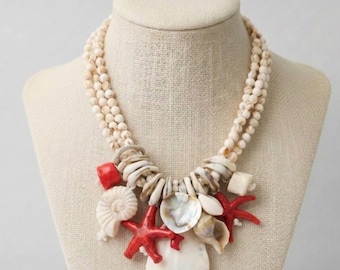May include: A multi-strand necklace featuring cream-colored beads and a variety of seashells. The necklace is adorned with red starfish, white shells, and a large, white shell pendant, creating a beachy aesthetic.