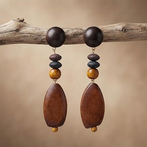 May include: A pair of dangle earrings featuring a large dark brown wooden sphere at the top, followed by a small dark brown bead, a textured black bead, a smooth mustard yellow bead, and a large, flat, teardrop-shaped brown wooden pendant.