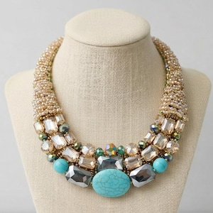 May include: A statement necklace with a multi-layered design. The necklace features large, faceted, clear and grey stones, turquoise beads, and smaller iridescent gems. The necklace is set on a beaded gold-toned base and displayed on a mannequin.