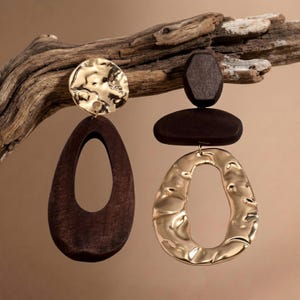 May include: A pair of mismatched earrings featuring dark brown wood and gold-toned metal. One earring has a circular hammered gold top with a large dark brown wooden teardrop pendant. The other has a dark brown wooden top with a gold teardrop.