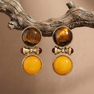 May include: A pair of drop earrings featuring a round, brown tiger's eye stone at the top and a round, opaque yellow stone at the bottom. The two stones are connected by a metallic gold-toned bar with small, round, red accents on each side.