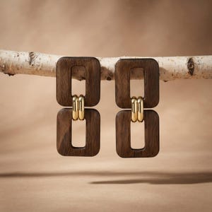 May include: A pair of drop earrings featuring two dark brown rectangular wooden frames connected by two polished gold-tone metal rings. The earrings are suspended from a light-colored birch branch, showcasing a geometric design with wood and metal.