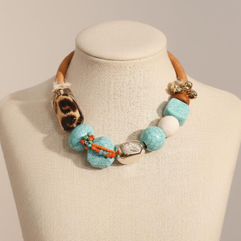 May include: A statement necklace featuring a tan leather cord with a variety of beads. The beads include turquoise, white, silver, and leopard print. The necklace is displayed on a mannequin.