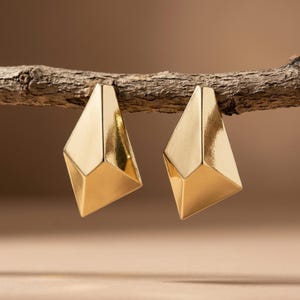 Puede incluir: Un par de pendientes dorados con una forma geométrica, multifacética y angular. Cada pendiente presenta una superficie lisa y reflectante con bordes afilados y una base puntiaguda, suspendidos de una rama de madera horizontal y texturizada.