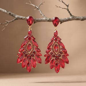 May include: A pair of chandelier earrings featuring numerous faceted red crystals set in gold-toned metal. Each earring has a teardrop-shaped top stone connected to a large, fan-shaped drop composed of multiple marquise and round red crystals.