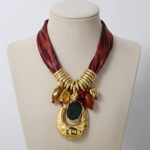 Puede incluir: Un collar llamativo con una banda roja y dorada similar a una cinta. El collar presenta detalles metálicos dorados y un colgante grande de oro martillado con un centro verde oscuro. Varias cuentas de color ámbar y rojo cuelgan del colgante.