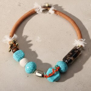 May include: A handcrafted necklace featuring a tan cord, turquoise beads, and a clear, patterned cylinder. Accents include a gold-toned floral charm, a silver-toned oval bead, and a red beaded strand. The necklace has a unique, bohemian style.