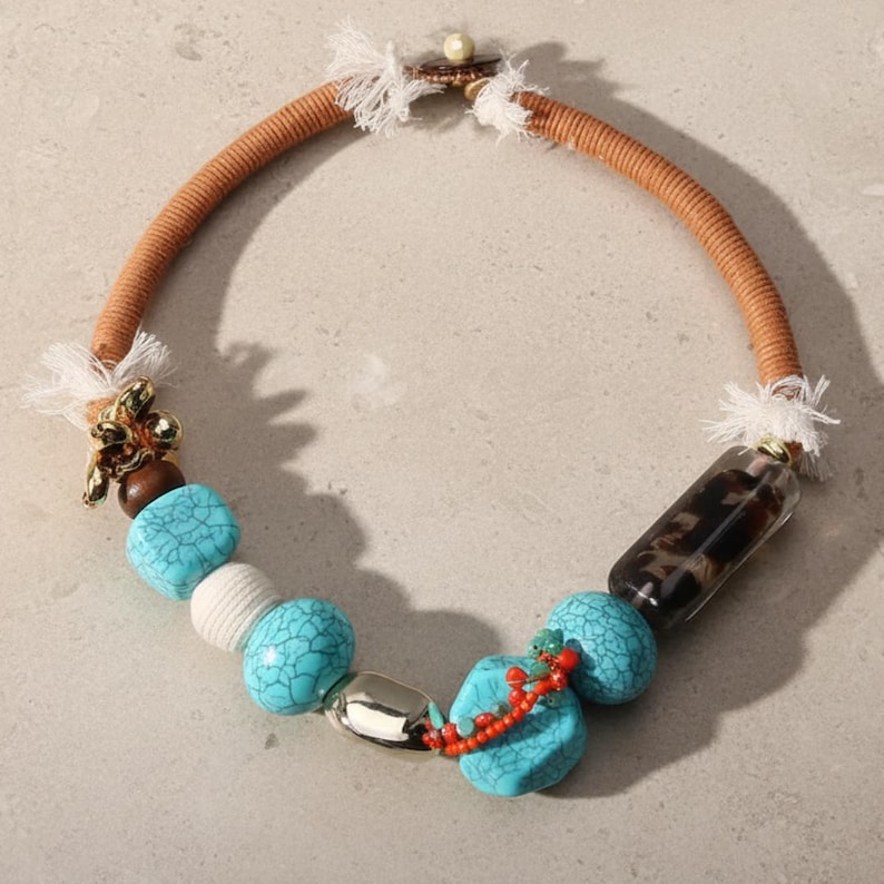 May include: A handcrafted necklace featuring a tan cord, turquoise beads, and a clear, patterned cylinder. Accents include a gold-toned floral charm, a silver-toned oval bead, and a red beaded strand. The necklace has a unique, bohemian style.