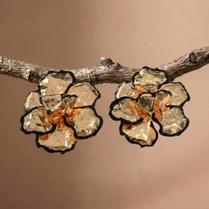 Könnte beinhalten: Ein Paar goldfarbene, blumenförmige Ohrringe mit einer gehämmerten, reflektierenden Oberflächenstruktur und dunklen, kontrastierenden Rändern. Die Blütenblätter haben ein organisches, geschichtetes Aussehen für einen dreidimensionalen Effekt.