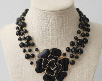 May include: A multi-strand necklace featuring glossy black beads of varying sizes strung on gold-tone metal links. The centerpiece is a large, layered black enamel flower with gold-tone edges, creating a bold and structured floral design.