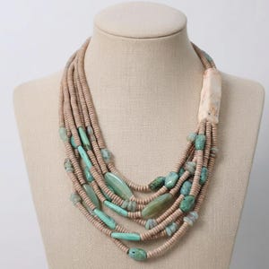 May include: A multi-strand necklace featuring light brown beads and turquoise-colored stones. A large, rectangular, off-white stone is at the top. The necklace is displayed on a mannequin.