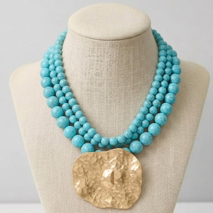 May include: A turquoise beaded necklace with a large, hammered gold-tone pendant. The necklace features multiple strands of round, light blue beads. The pendant has a textured surface and a geometric design.