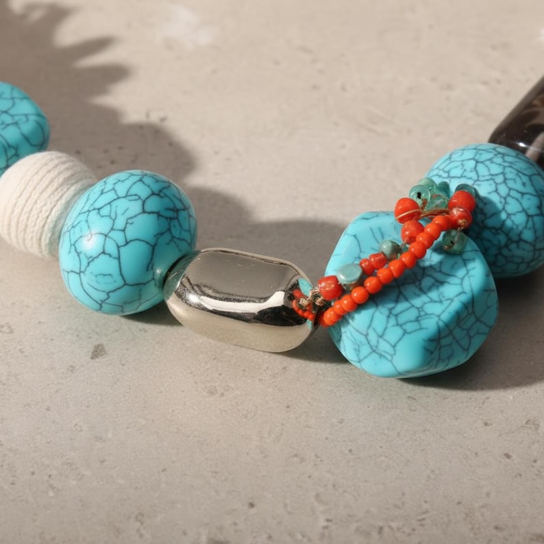 May include: Close-up of a necklace featuring large, turquoise-colored beads with a crackle effect. The necklace also includes a silver-colored, oval-shaped bead and small orange and green beads.