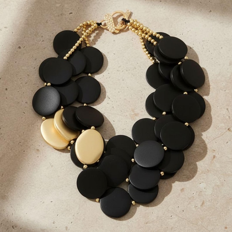 Pode incluir: Um colar marcante com pingentes circulares pretos e dourados. O colar tem um fecho dourado e detalhes de contas douradas. O design incorpora elementos pretos foscos e dourados brilhantes, criando um contraste ousado.