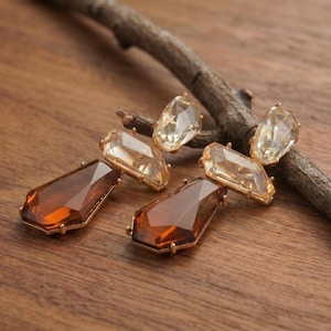 May include: A pair of drop earrings featuring faceted, geometric crystals in a gradient of amber and light champagne tones. Each earring consists of a smaller, lighter-coloured top crystal connected to a larger, darker amber-coloured bottom crystal.