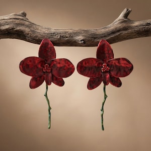 Może przedstawiać: Para kolczyków w kształcie ciemnoczerwonych kwiatów orchidei o fakturze młotkowanego metalu. Każdy kwiat posiada w środku skupisko trzech małych czerwonych kryształków oraz cienką, zieloną, teksturowaną łodygę skierowaną ku dołowi.