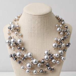 May include: A multi-strand necklace featuring large, round, gray faux pearls and smaller, faceted, dark gray beads. The necklace also includes small gold-colored beads and is displayed on a beige mannequin.