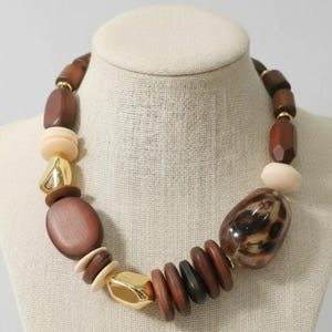 Puede incluir: Collar llamativo con una mezcla de cuentas de madera en tonos marrón, crema y negro. Acentuado con metal dorado y una gran cuenta de resina con estampado de leopardo. El collar se exhibe en un maniquí de tono neutro.