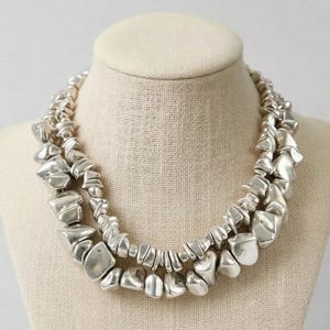 May include: A silver-toned necklace with two strands of irregularly shaped, polished metal beads. The necklace is displayed on a neutral-toned mannequin bust, showcasing its layered design and reflective surface.