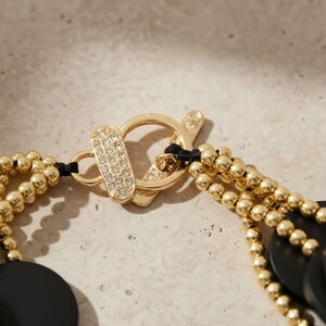 Pode incluir: Close-up de um colar dourado com fecho de altern&acirc;ncia. O fecho apresenta uma sec&ccedil;&atilde;o retangular incrustada com pequenos cristais transparentes. O colar tem v&aacute;rios fios de pequenas contas douradas e pingentes circulares pretos.