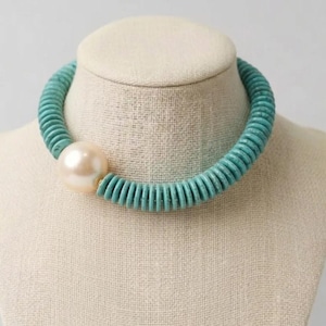 May include: A turquoise beaded choker necklace with a large, off-white pearl accent. The necklace is displayed on a mannequin bust. The beads are small and round, creating a textured, chunky look. The pearl is smooth and spherical.